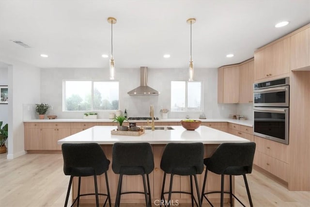 kitchen featuring appliances with stainless steel finishes, light brown cabinetry, a kitchen island with sink, and wall chimney range hood