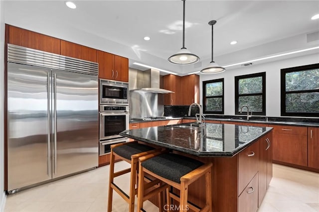 kitchen featuring a breakfast bar area, hanging light fixtures, a kitchen island with sink, built in appliances, and wall chimney range hood