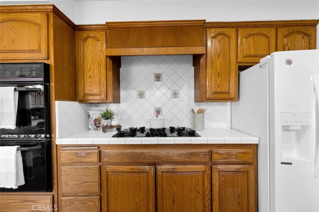 kitchen featuring wall chimney range hood, tile counters, decorative backsplash, and black appliances