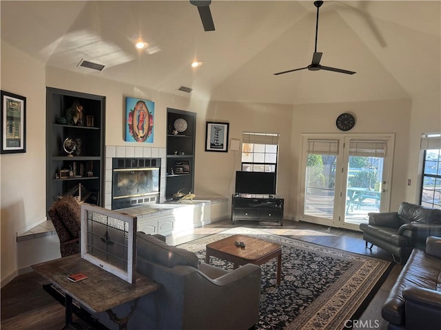 living area with built in shelves, lofted ceiling, visible vents, a tiled fireplace, and a ceiling fan