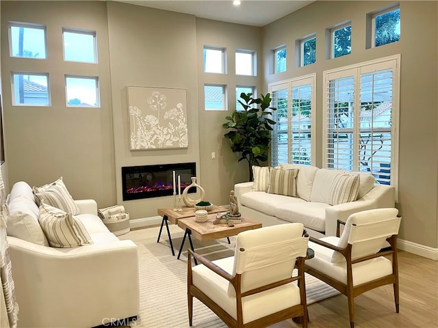 living room featuring light wood-type flooring, a high ceiling, baseboards, and a glass covered fireplace