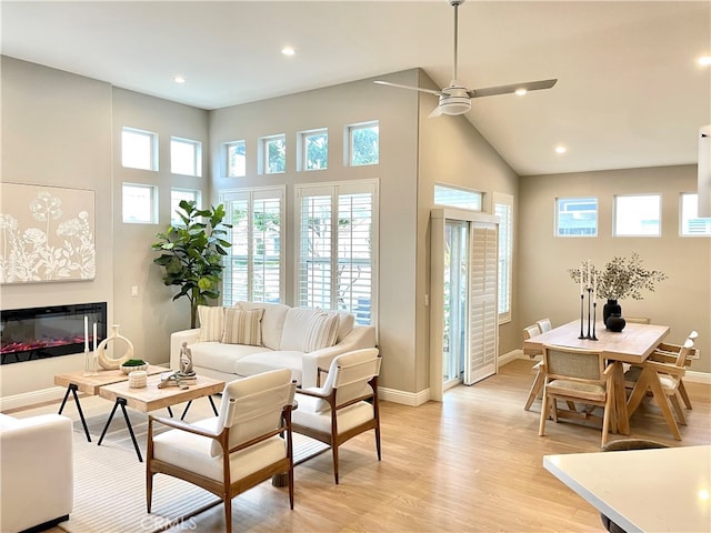 living area with light wood-style flooring, a high ceiling, a ceiling fan, baseboards, and a glass covered fireplace