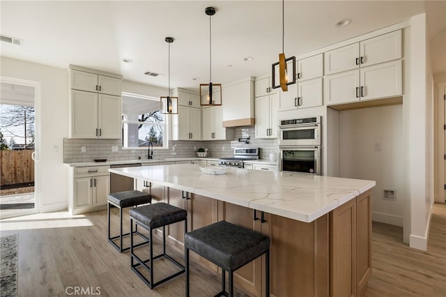 kitchen with pendant lighting, appliances with stainless steel finishes, light stone counters, custom range hood, and a kitchen island