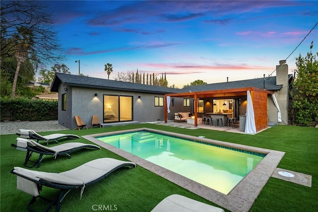 back house at dusk with a patio, an outdoor bar, and a lawn