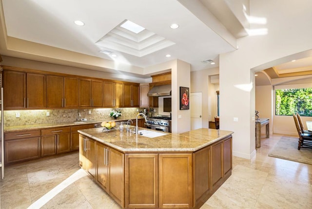 kitchen with decorative backsplash, light stone counters, a tray ceiling, wall chimney range hood, and a spacious island