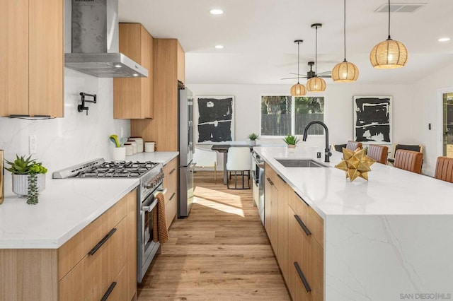 kitchen featuring sink, appliances with stainless steel finishes, a kitchen island with sink, decorative light fixtures, and wall chimney exhaust hood