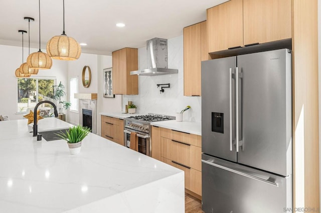 kitchen featuring wall chimney exhaust hood, light stone counters, hanging light fixtures, light brown cabinets, and premium appliances