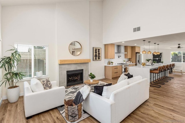 living room with sink, ceiling fan, a high ceiling, a tiled fireplace, and light wood-type flooring