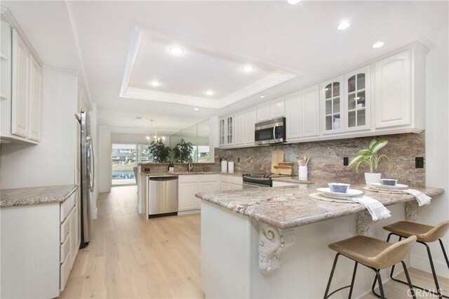 kitchen with a raised ceiling, appliances with stainless steel finishes, glass insert cabinets, white cabinets, and a peninsula