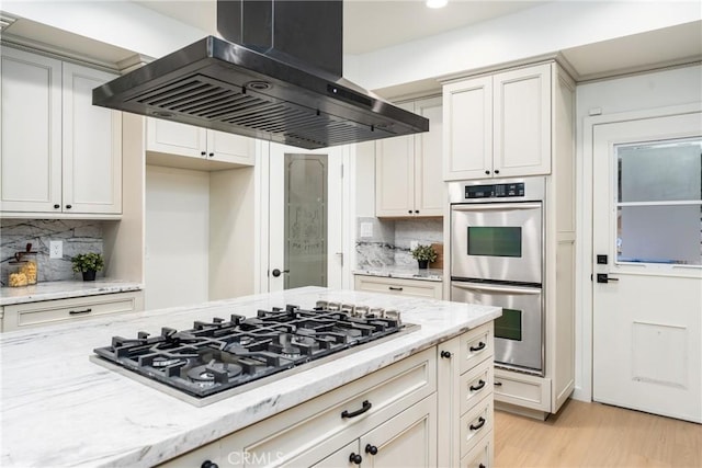 kitchen featuring stainless steel appliances, tasteful backsplash, light stone countertops, island exhaust hood, and light wood-type flooring