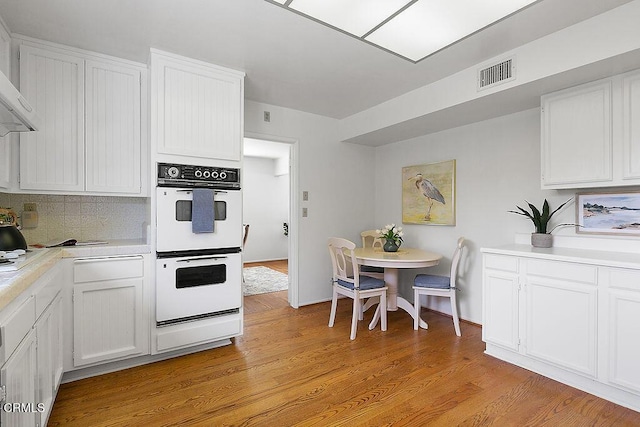 kitchen with wall chimney exhaust hood, white double oven, light wood-type flooring, and white cabinets