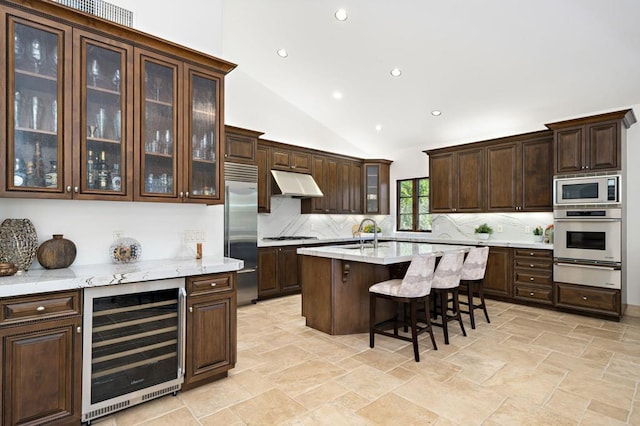 kitchen with beverage cooler, a kitchen island with sink, built in appliances, and dark brown cabinets