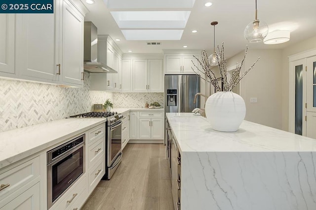 kitchen with appliances with stainless steel finishes, decorative light fixtures, white cabinetry, light stone counters, and wall chimney exhaust hood