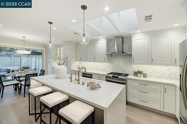 kitchen featuring pendant lighting, wall chimney range hood, sink, stainless steel appliances, and a center island with sink