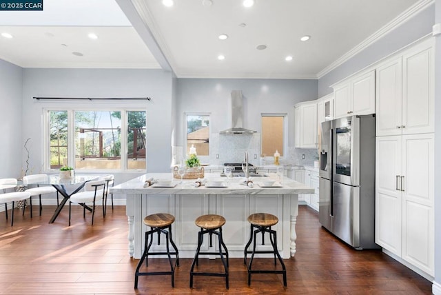 kitchen with wall chimney range hood, a breakfast bar area, a kitchen island with sink, white cabinetry, and stainless steel refrigerator with ice dispenser