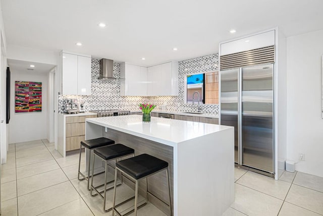 kitchen with wall chimney range hood, a center island, stainless steel built in refrigerator, white cabinets, and a kitchen bar