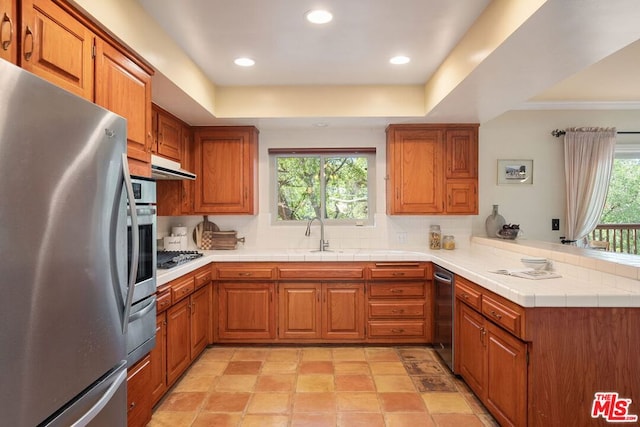 kitchen with tasteful backsplash, tile countertops, a tray ceiling, kitchen peninsula, and stainless steel appliances