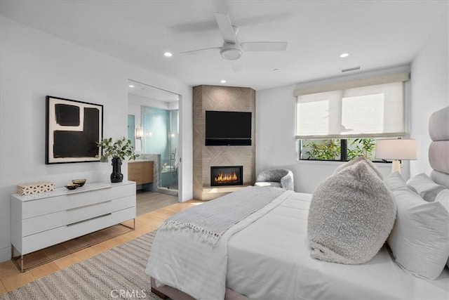 bedroom with ensuite bathroom, a large fireplace, ceiling fan, and light wood-type flooring