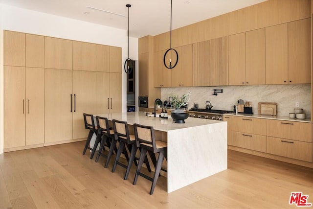 kitchen featuring a breakfast bar, a kitchen island with sink, light brown cabinets, decorative backsplash, and light wood-type flooring