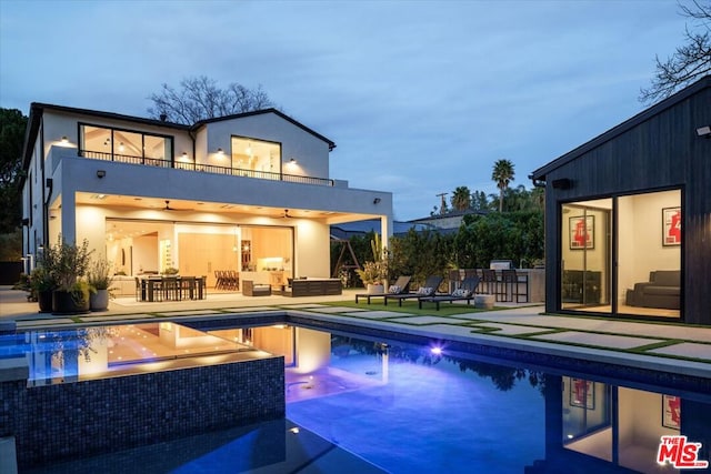 back house at dusk with a balcony, a swimming pool with hot tub, outdoor lounge area, and a patio area