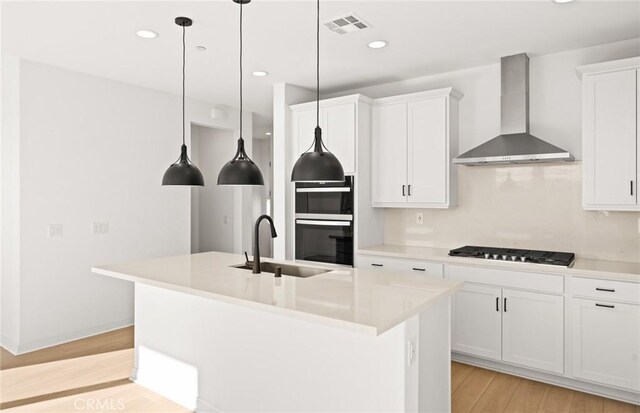 kitchen with wall chimney range hood, sink, a kitchen island with sink, white cabinetry, and black gas cooktop