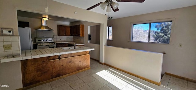 kitchen with wall chimney range hood, stainless steel stove, fridge, tile counters, and kitchen peninsula