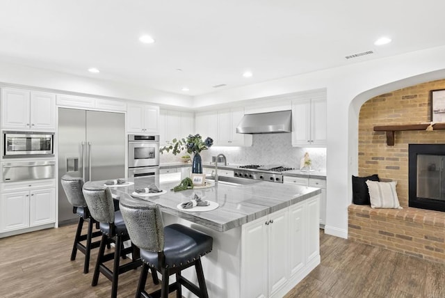 kitchen featuring an island with sink, sink, white cabinets, built in appliances, and wall chimney exhaust hood