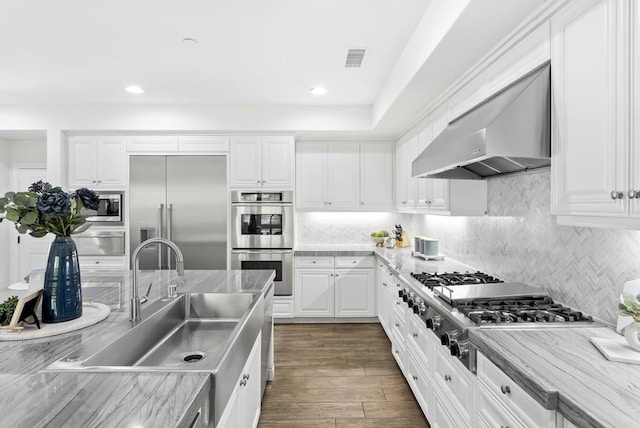 kitchen featuring tasteful backsplash, white cabinetry, sink, built in appliances, and wall chimney range hood