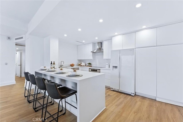 kitchen featuring a breakfast bar area, white cabinets, white refrigerator with ice dispenser, light wood-type flooring, and wall chimney exhaust hood