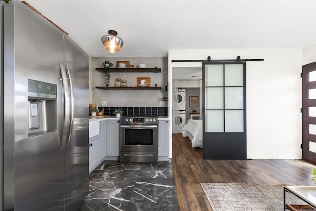 kitchen with appliances with stainless steel finishes, gray cabinetry, backsplash, stacked washer / dryer, and a barn door