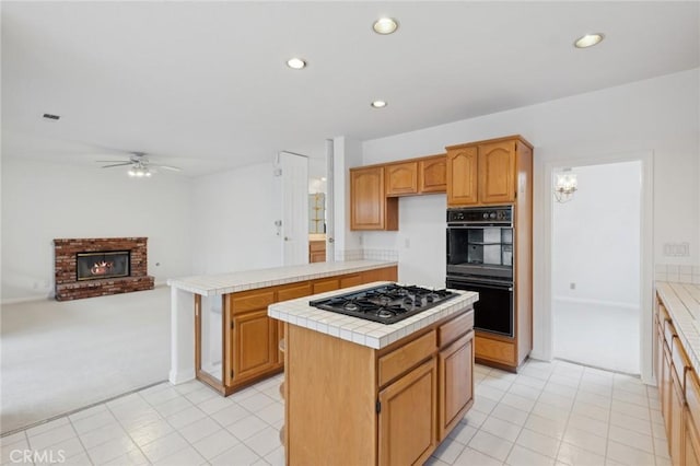 kitchen featuring tile countertops, a center island, ceiling fan, black appliances, and a brick fireplace