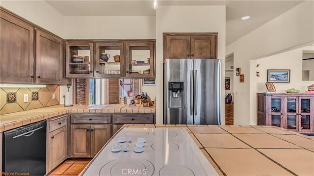 kitchen featuring tile countertops, stainless steel refrigerator with ice dispenser, backsplash, dishwasher, and glass insert cabinets