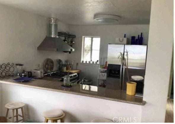 kitchen featuring stainless steel fridge with ice dispenser, a kitchen bar, stove, kitchen peninsula, and wall chimney exhaust hood