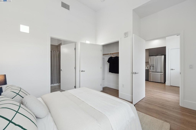 bedroom with connected bathroom, a walk in closet, light wood-type flooring, stainless steel fridge, and a high ceiling