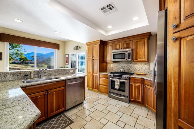 kitchen with sink, appliances with stainless steel finishes, backsplash, light stone counters, and a tray ceiling