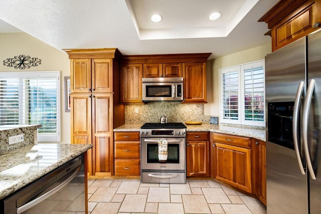 kitchen with light stone counters, appliances with stainless steel finishes, a tray ceiling, a wealth of natural light, and backsplash