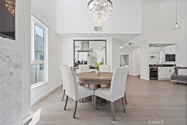 dining space with sink, a towering ceiling, a notable chandelier, beverage cooler, and light wood-type flooring