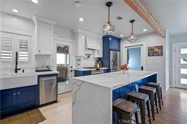 kitchen with white cabinetry, a center island, stainless steel appliances, blue cabinetry, and wall chimney range hood