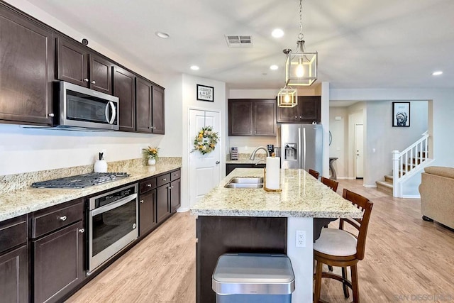 kitchen with sink, appliances with stainless steel finishes, dark brown cabinetry, light hardwood / wood-style floors, and a center island with sink