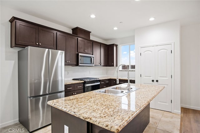 kitchen featuring appliances with stainless steel finishes, an island with sink, sink, light stone counters, and dark brown cabinets