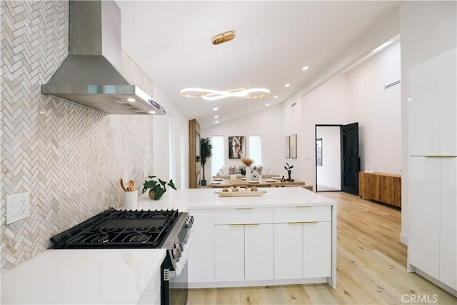 kitchen with lofted ceiling, stainless steel gas range, white cabinetry, decorative light fixtures, and wall chimney exhaust hood