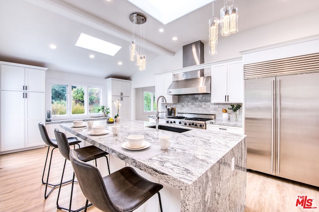 kitchen featuring stainless steel built in fridge, an island with sink, wall chimney range hood, light stone countertops, and white cabinets