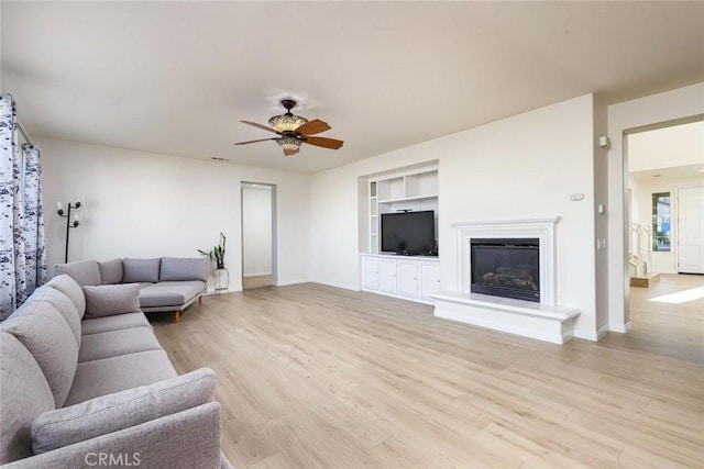 living room with light wood finished floors, baseboards, built in features, a ceiling fan, and a glass covered fireplace