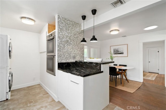 kitchen featuring stacked washer and clothes dryer, white cabinets, decorative light fixtures, kitchen peninsula, and stainless steel double oven