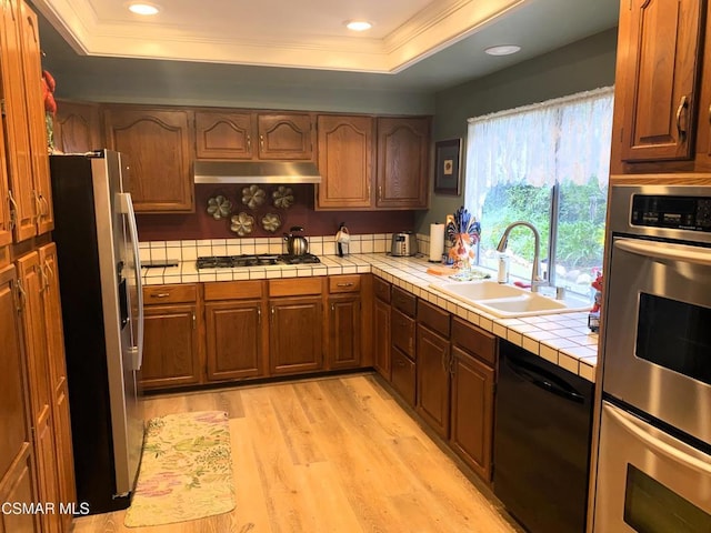 kitchen with tile countertops, a raised ceiling, sink, stainless steel appliances, and light hardwood / wood-style flooring