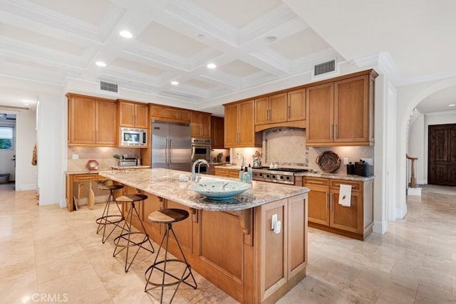 kitchen featuring a breakfast bar, built in appliances, a center island with sink, beam ceiling, and light stone countertops