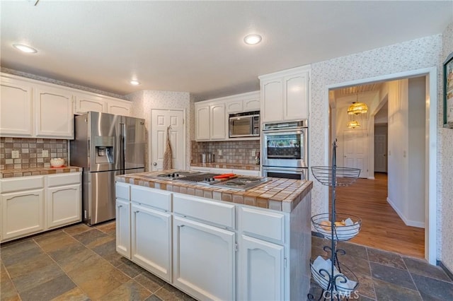 kitchen with tasteful backsplash, white cabinets, appliances with stainless steel finishes, tile counters, and wallpapered walls