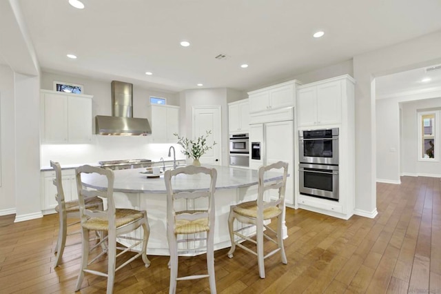 kitchen with double oven, white cabinets, dark hardwood / wood-style flooring, a kitchen island with sink, and wall chimney exhaust hood