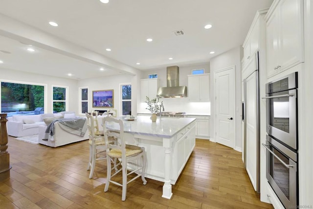 kitchen with wall chimney range hood, white cabinetry, a kitchen breakfast bar, an island with sink, and light wood-type flooring