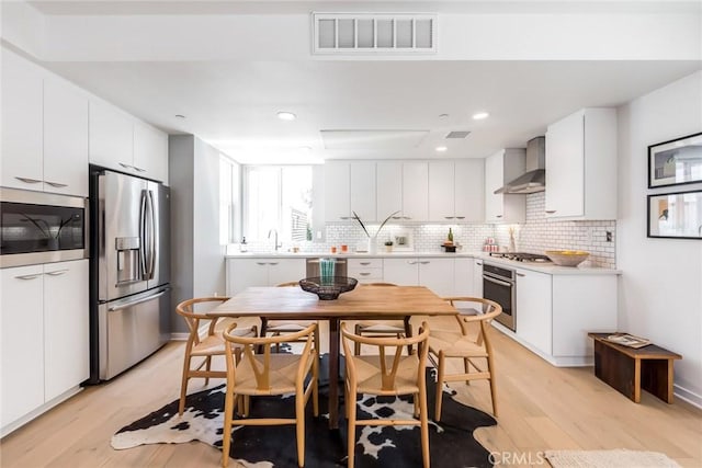 kitchen with white cabinetry, decorative backsplash, light hardwood / wood-style floors, stainless steel appliances, and wall chimney exhaust hood